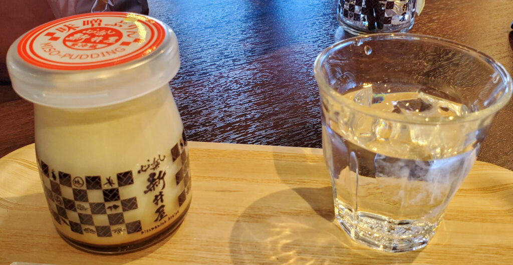 新井屋たぬまの杜本店の味噌ぷりん。市松模様のラベルが付いた瓶入りのプリンと水のグラス