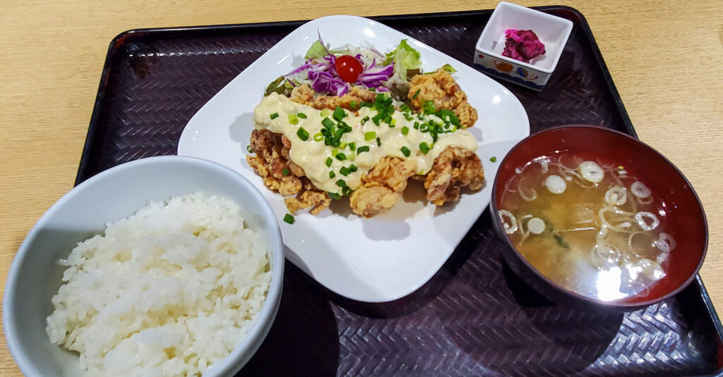 タルタルソースをかけた鶏の唐揚げ、ご飯、味噌汁、漬物が並ぶ定食
