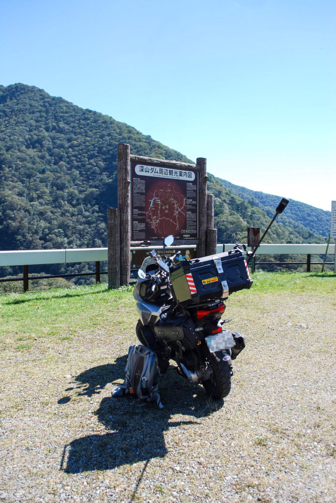深山ダム周辺観光案内図の前に駐車したHonda ADV160。背後には緑の山並みが広がる。