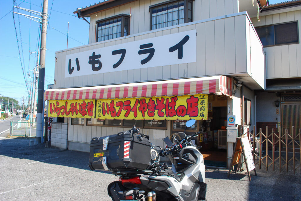 栃木県佐野市の江原商店。いもフライと焼きそばの看板が掲げられた店舗前に、ツーリングで訪れたADV160が停まっている。